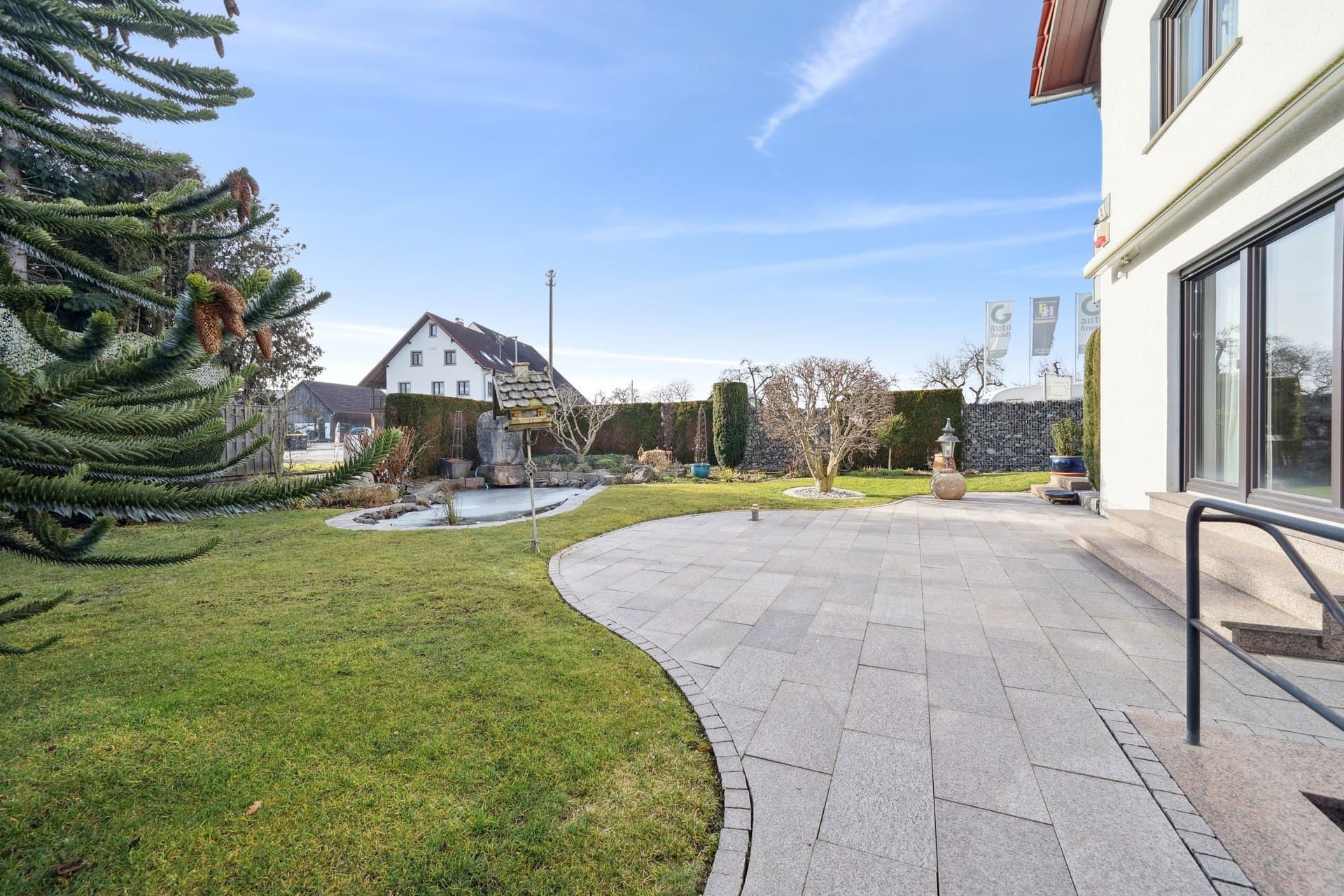 Gartenseite mit Rasenfläche, Araukarie und gepflasterter Terrasse unter blauem Himmel
